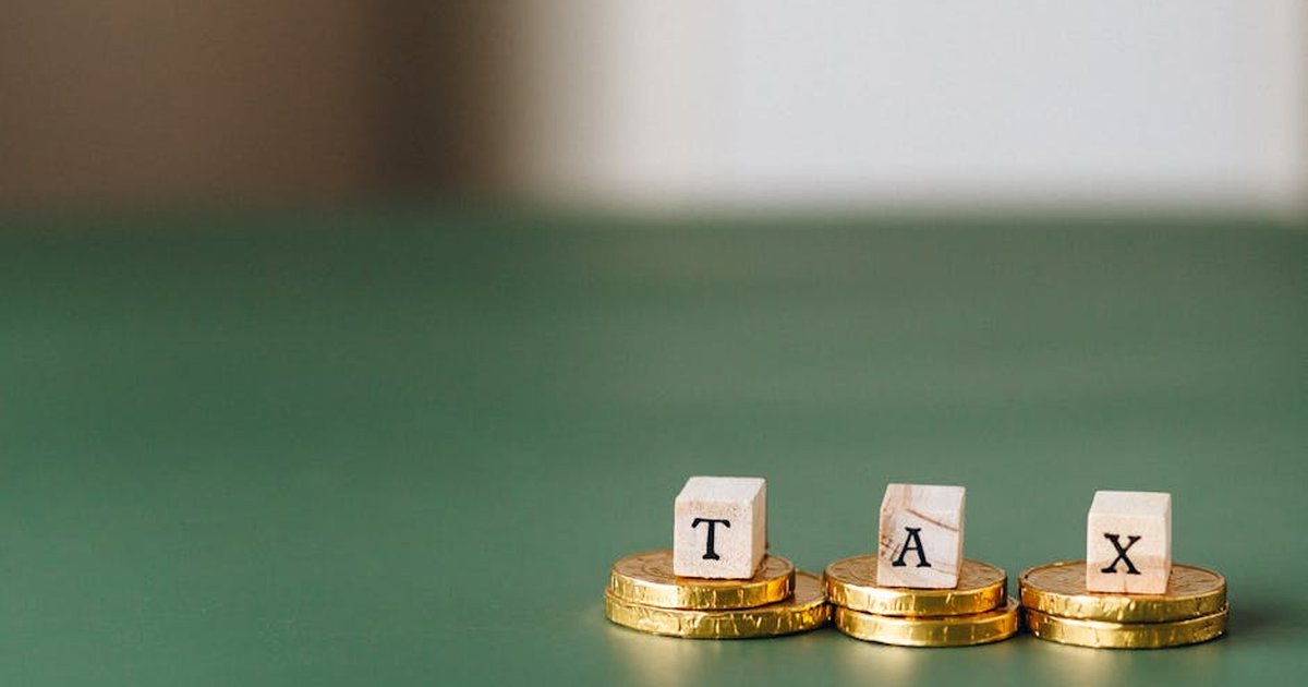 Stacked gold coins with wooden blocks spelling 'TAX' on a green backdrop.