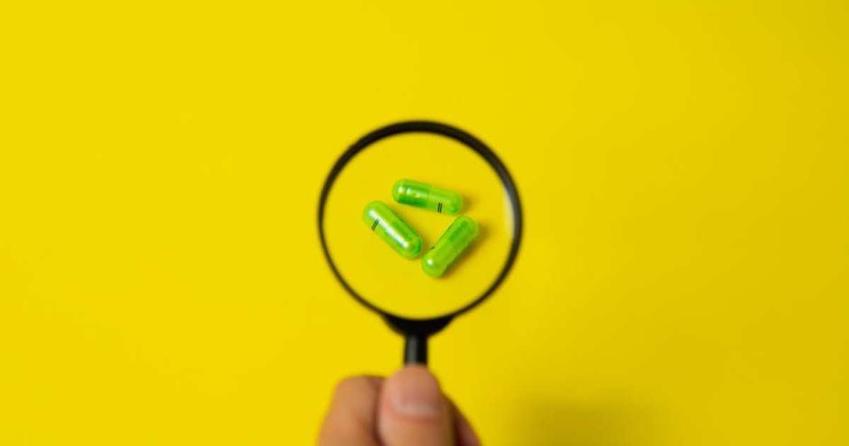 Top view of crop unrecognizable person holding magnifier over green pills placed on bright yellow surface in studio