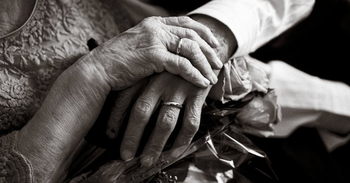 Black and white photo of an elderly couple holding hands with wedding rings, symbolizing love and companionship.