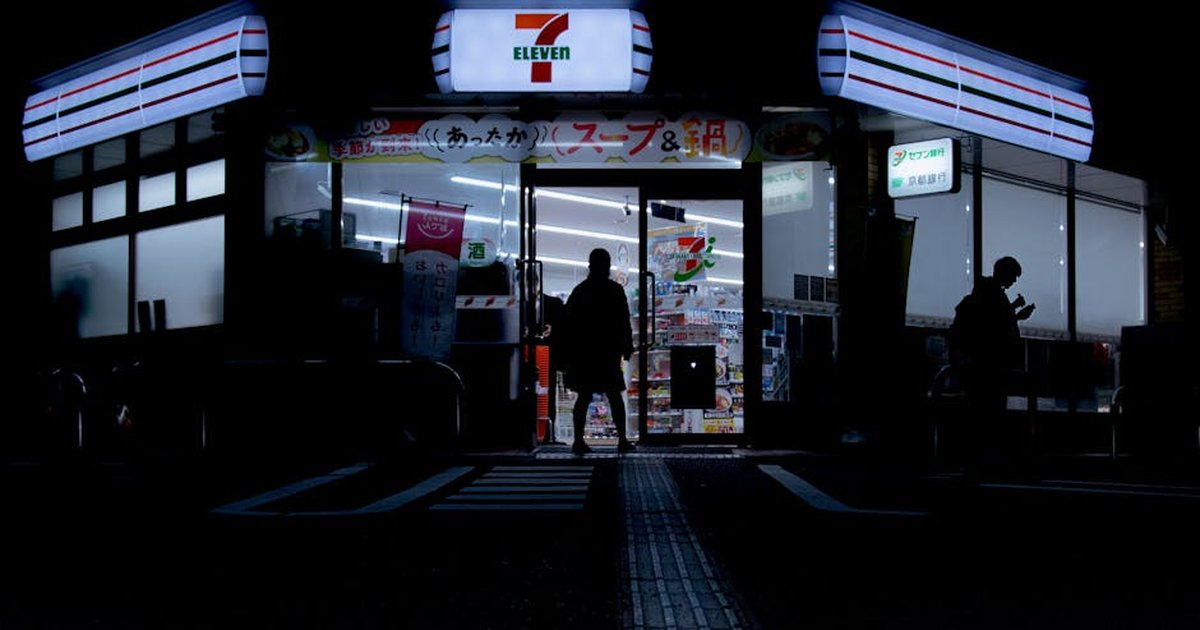 Silhouette of people entering a 7-Eleven store at night in Tokyo's urban setting.