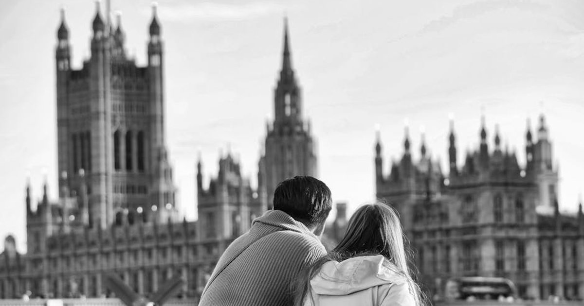 A couple sits together, admiring the iconic Westminster Palace in a black and white photograph.