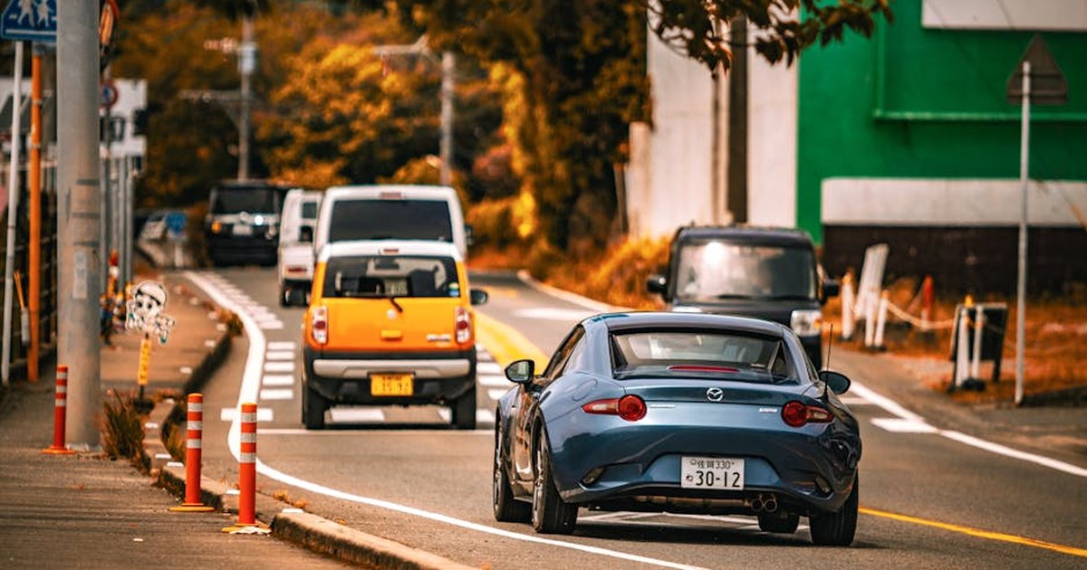 Mazda MX-5 car driving on an urban street surrounded by autumn foliage and city elements.