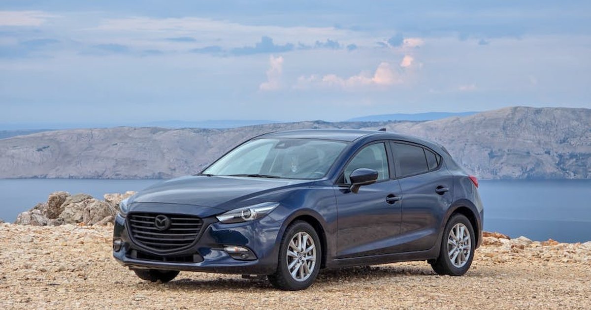 Blue sedan car parked on a rocky seashore with scenic mountainous backdrop under a cloudy sky.