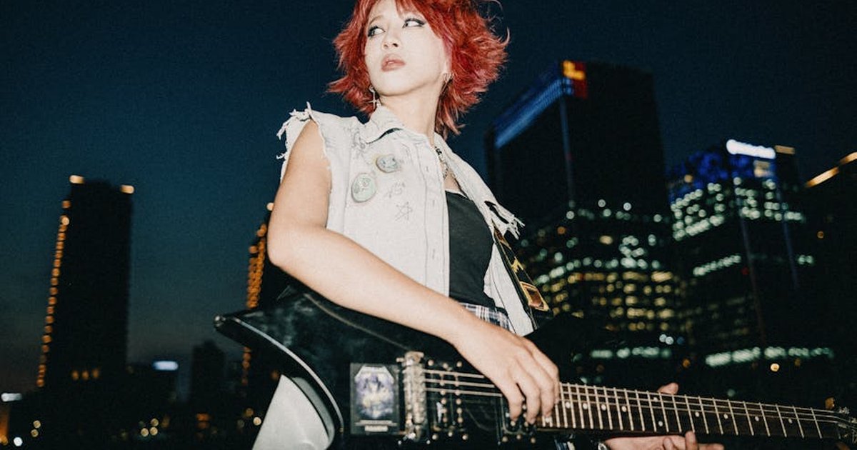 A woman with dyed hair plays an electric guitar against a city night skyline, showing urban rock vibes.