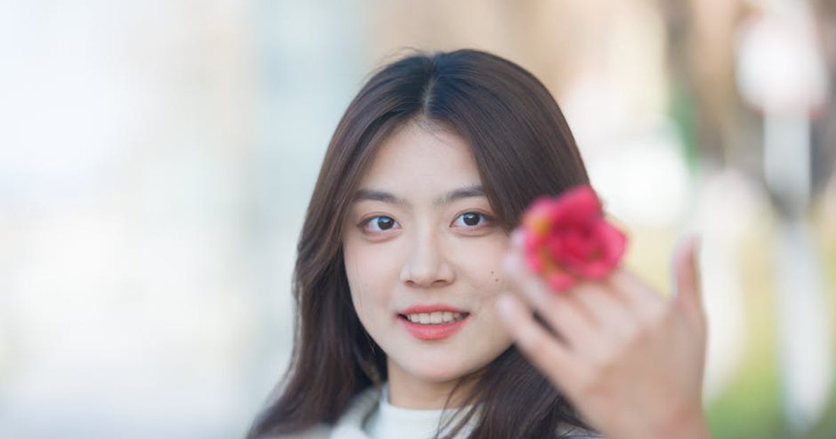A young woman smiling while holding a pink flower in a city environment.