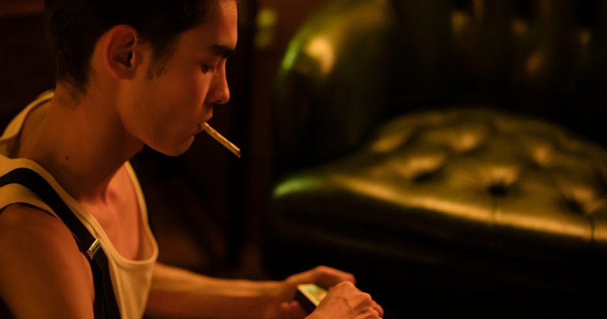 A young man sits indoors, smoking a cigarette in a dimly lit room.