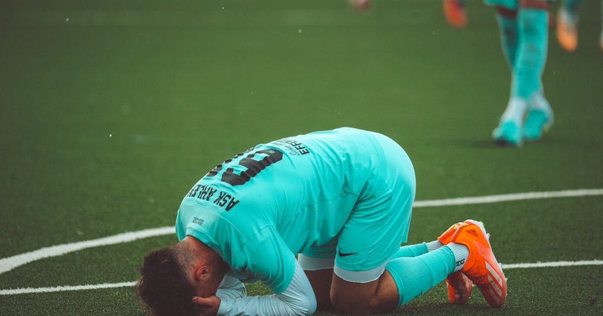 A soccer player kneels in despair on a green artificial turf sports field.