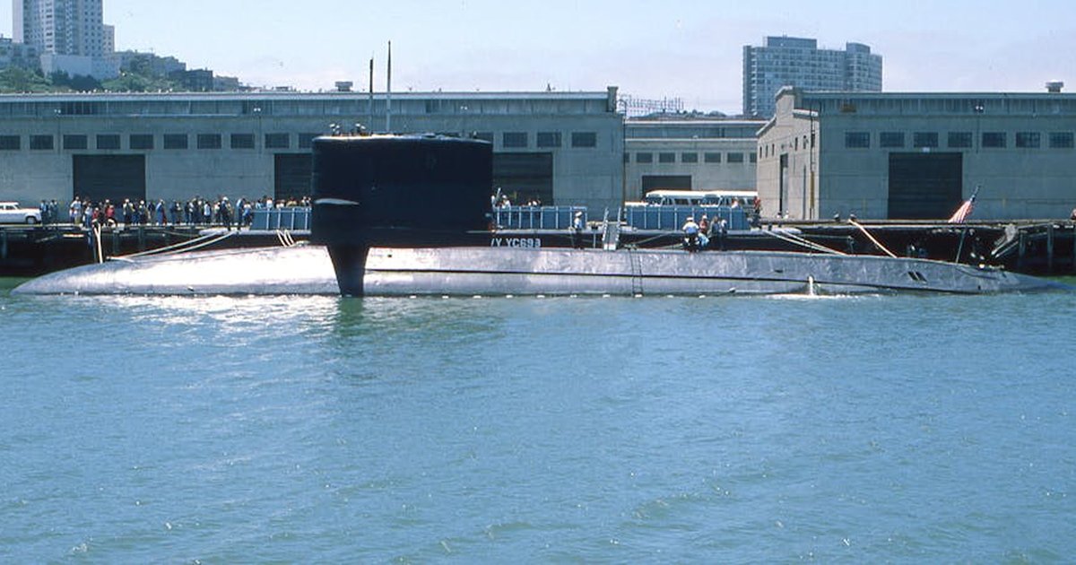A naval submarine docked at an urban harbor with people on shore.