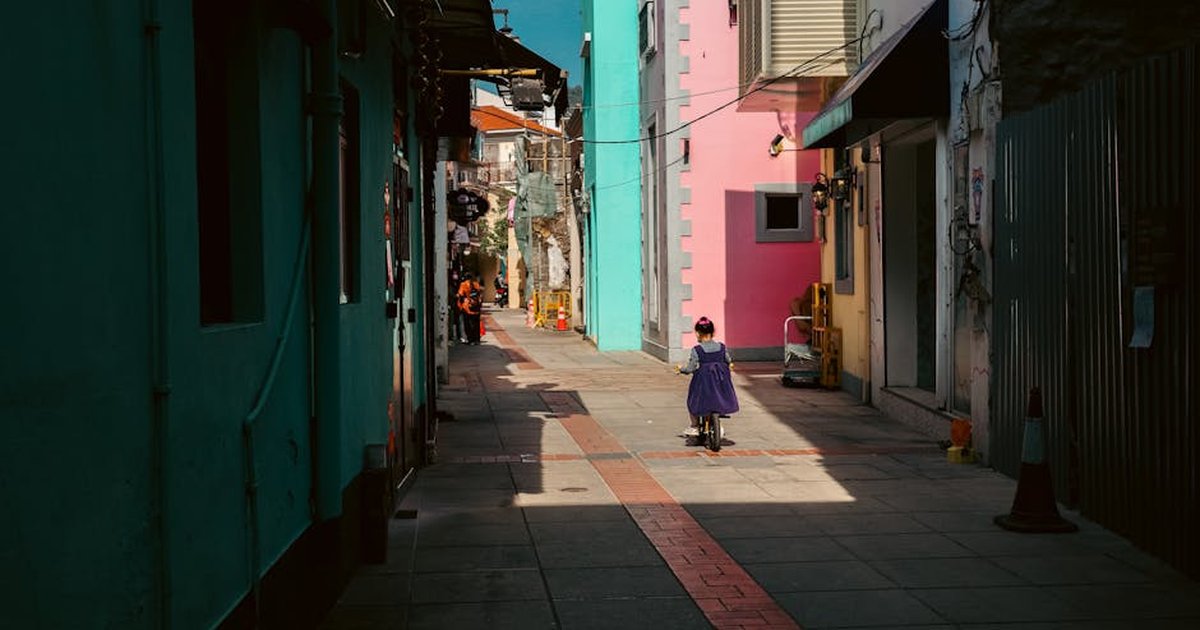 A child on a scooter explores a vibrant alleyway in Macau, filled with colorful buildings and warm sunlight.