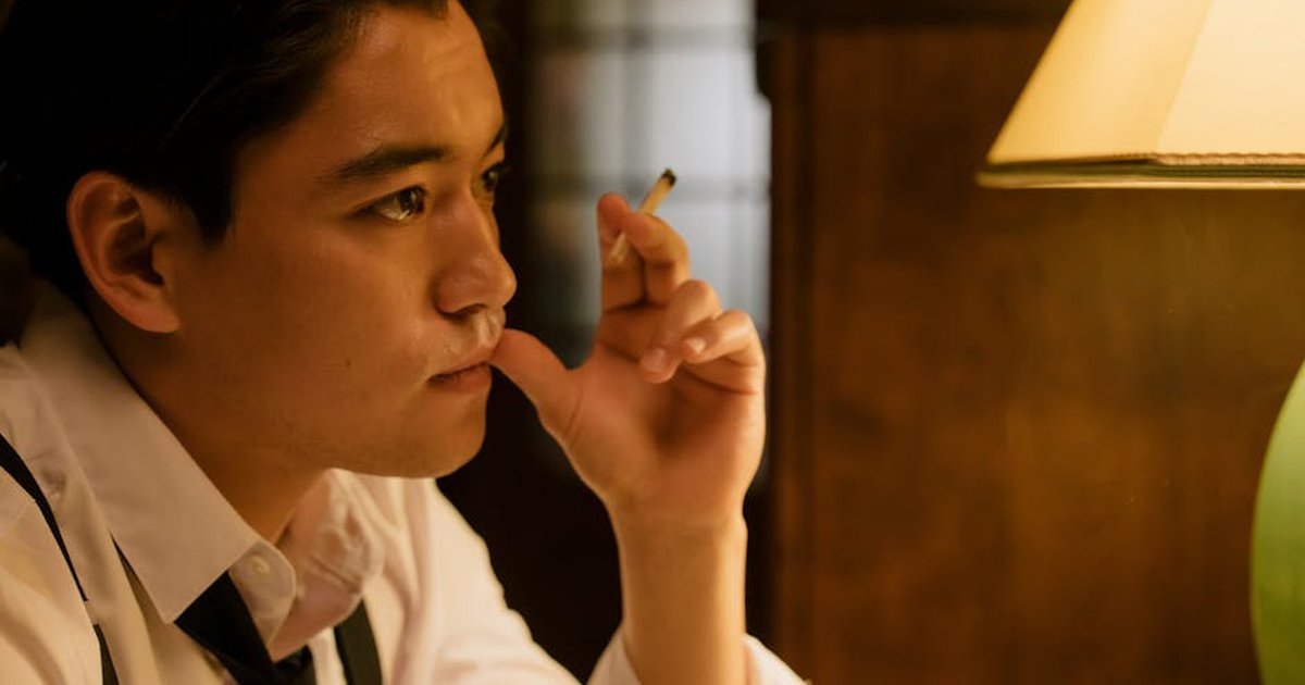 A thoughtful man with a cigarette, sitting indoors lit by a warm lamp.