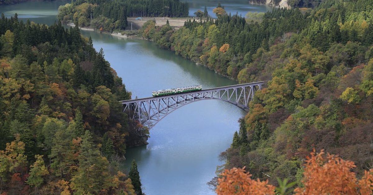 A scenic train traverses a bridge over a river, surrounded by vibrant autumn foliage.
