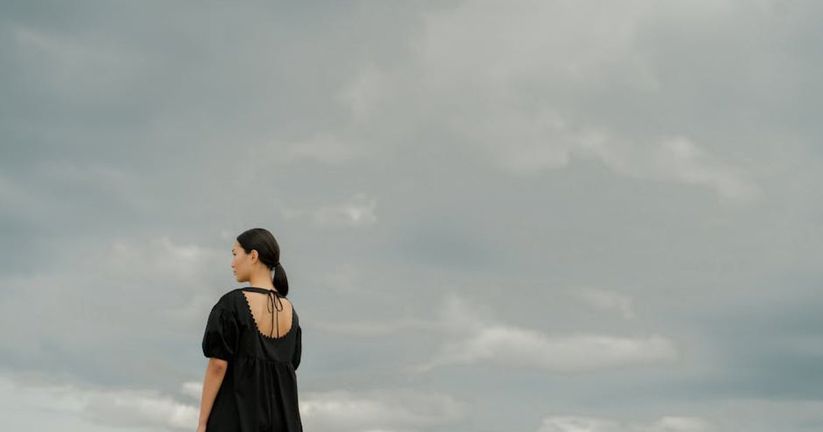 A woman in a black dress stands alone outdoors, gazing at a dramatic cloudy sky.