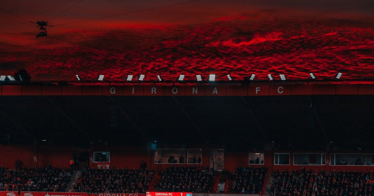 A stadium filled with fans under a dramatic red sunset sky during a Girona FC match.