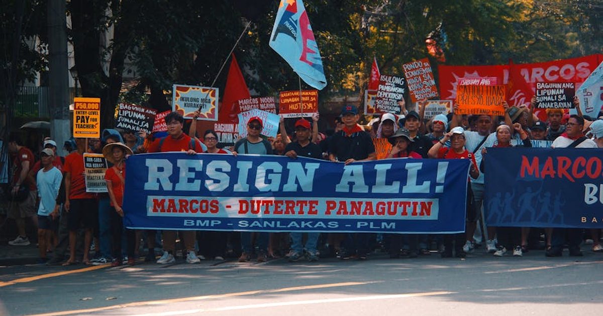 Crowd gathers in street protest holding signs demanding resignation with political messages.