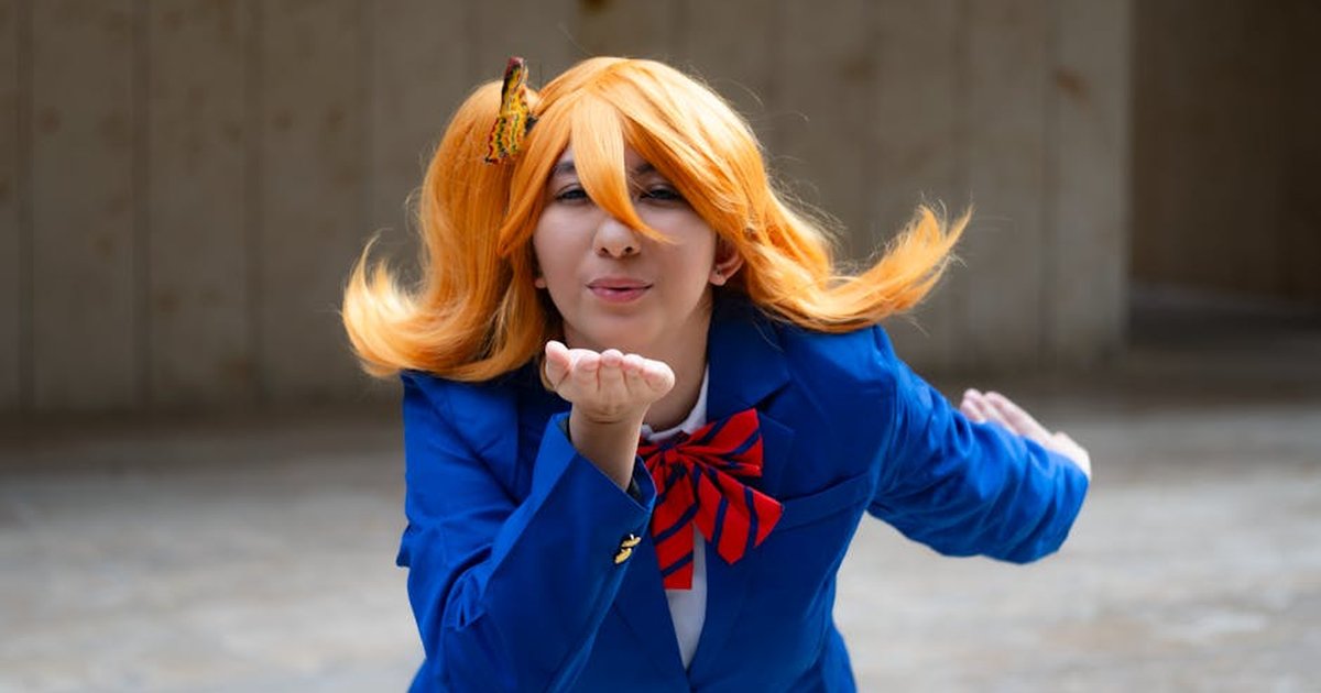 A vibrant cosplay portrait of a person in a blue suit and orange wig blowing a kiss outdoors.