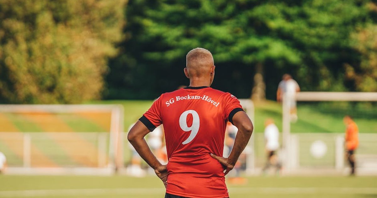 Back view of a soccer player in a red jersey on a green field during a game.