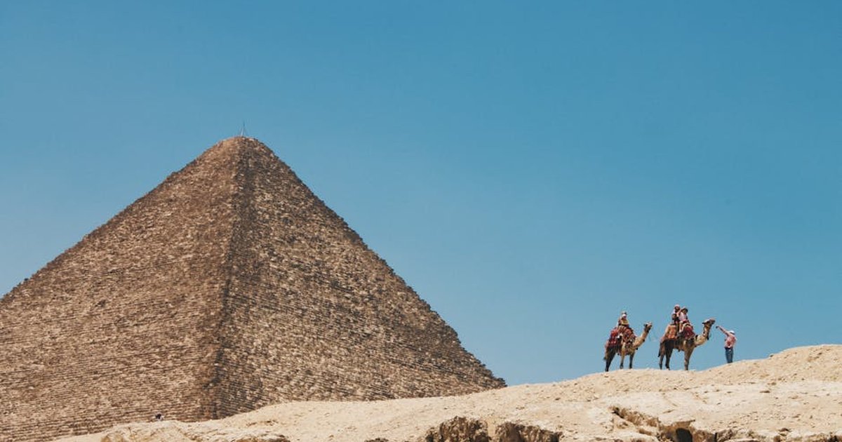 Camels and riders traverse the desert near the Great Pyramid of Giza under a blue sky.