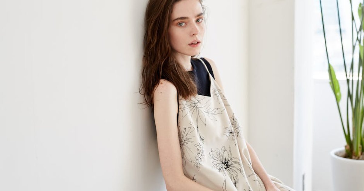 Caucasian woman in a floral dress leans against a white wall with indoor plants nearby.