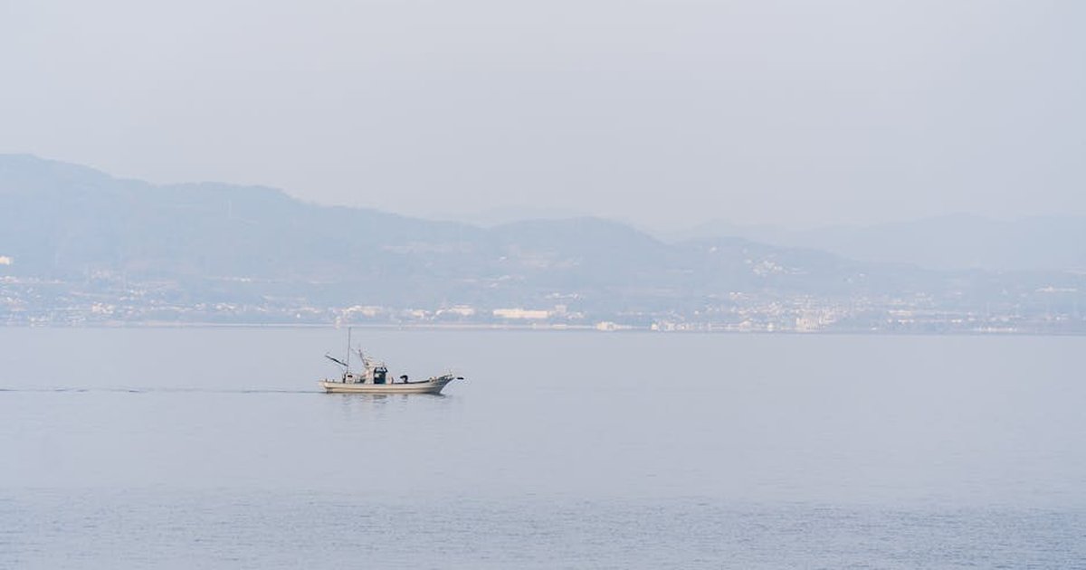 A fishing boat sails quietly across a calm and foggy sea with mountains in the background.