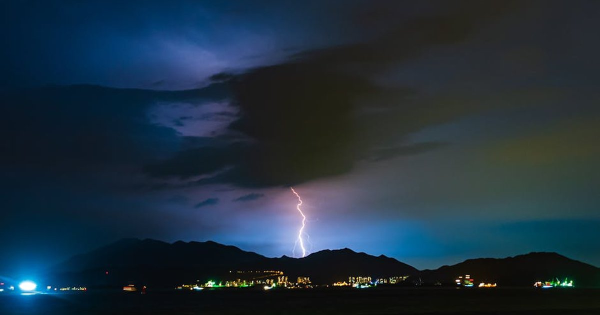 Nighttime lightning storm illuminating mountain silhouette with vibrant city lights below.