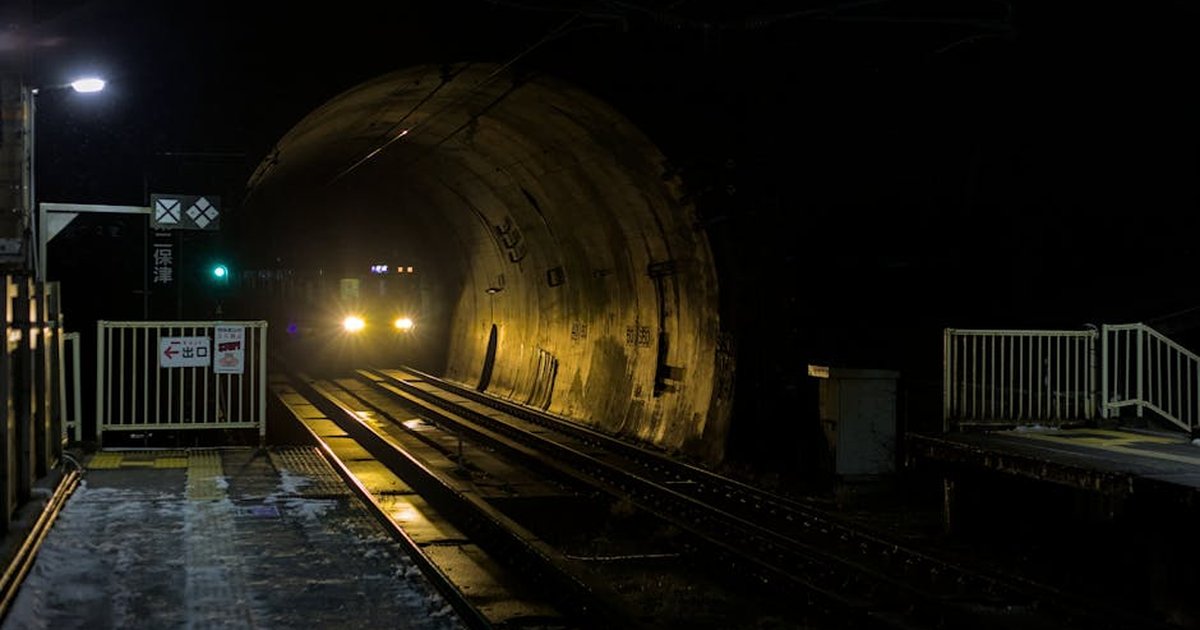 A train with headlights on approaches a snowy Hozukyō Station tunnel at night in winter, Kyoto, Japan.