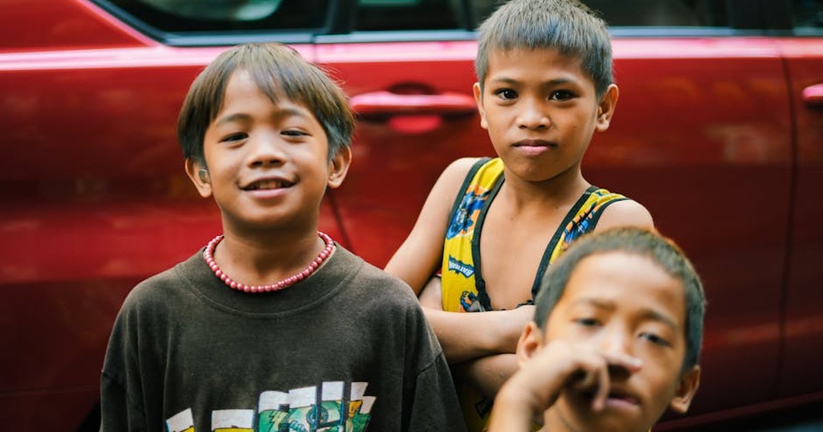 Three children standing together and smiling in front of a red car outdoors.