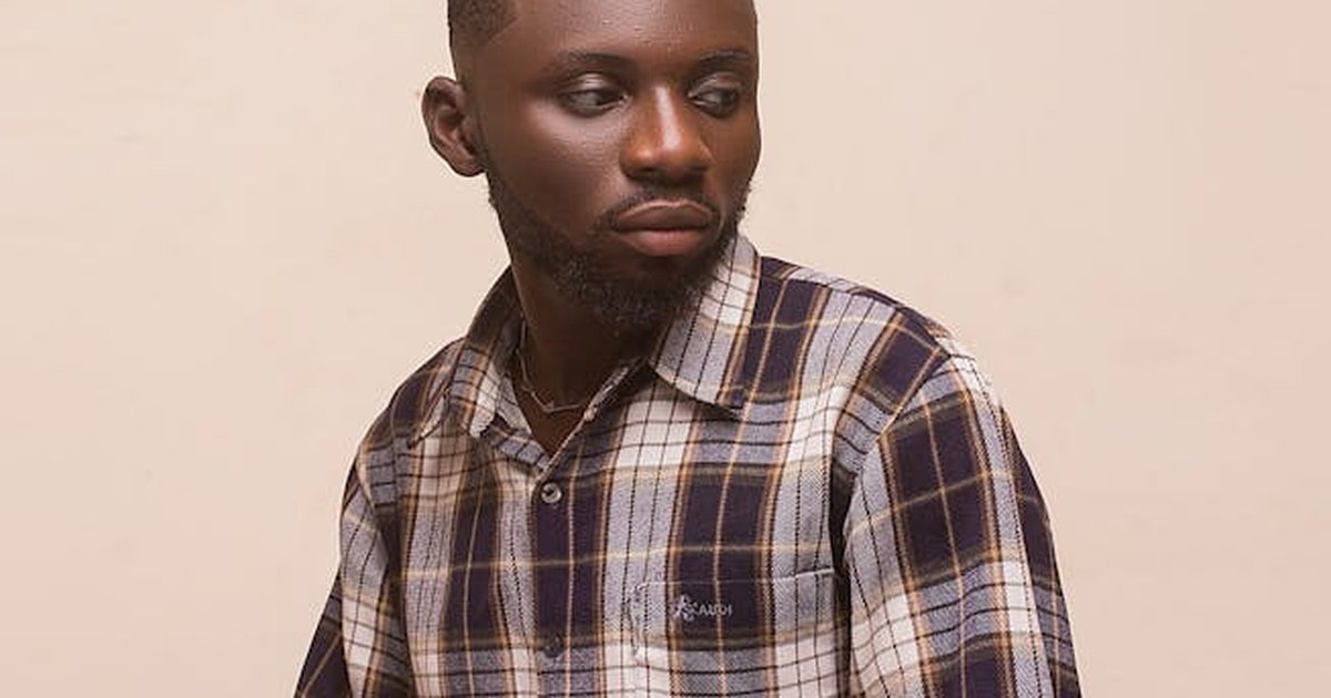 A young man wearing a plaid shirt sits, thoughtfully looking away.