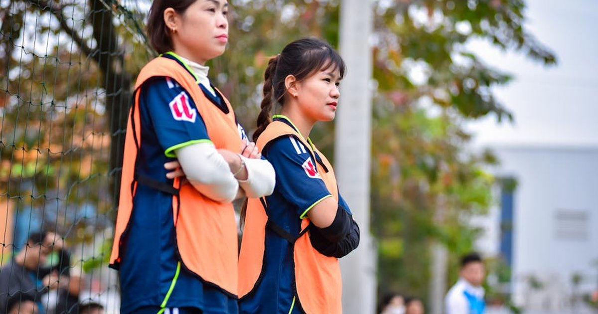 Two female football players in orange jerseys watch a game from the sideline in Hanoi, Vietnam.