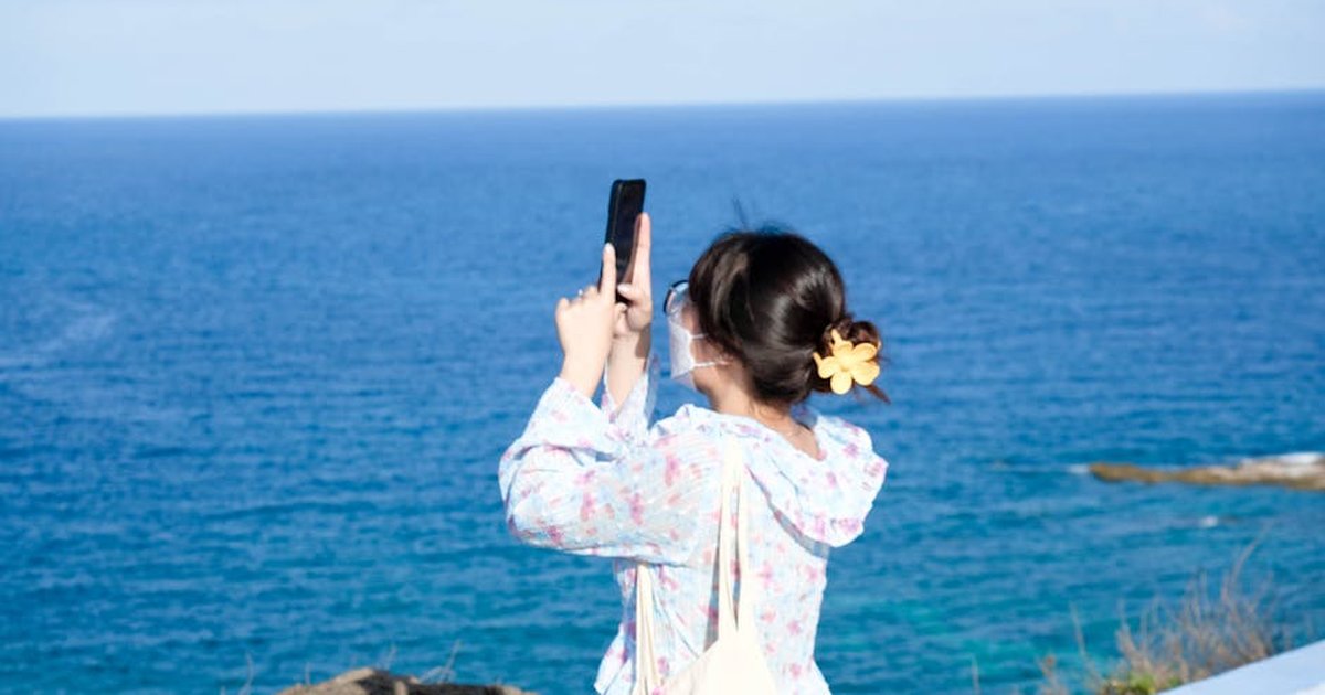 A woman taking a photo on a sunny day by the ocean with a smartphone.