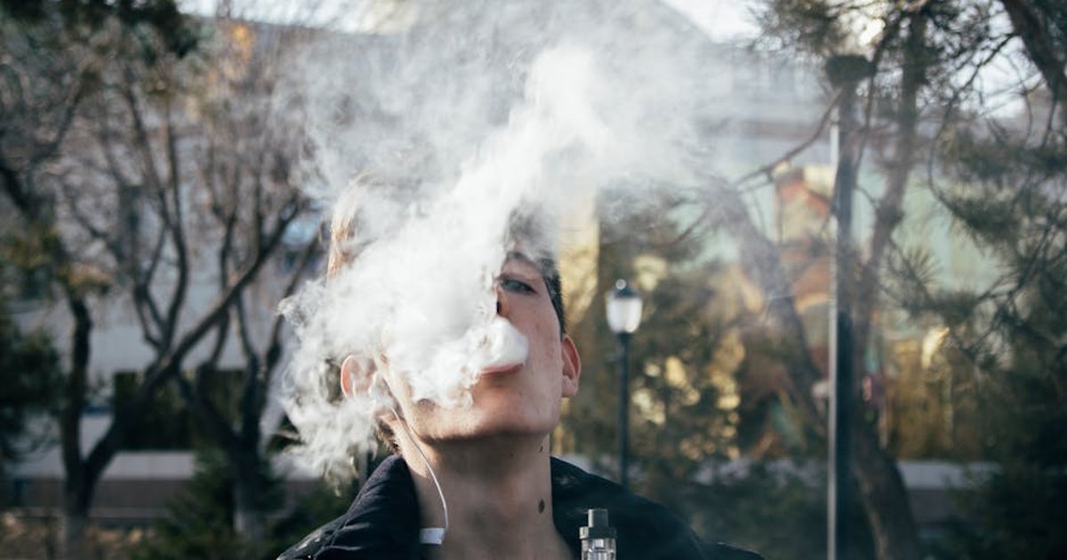 Portrait of a young man vaping in a park, releasing clouds of vapor on a clear day.