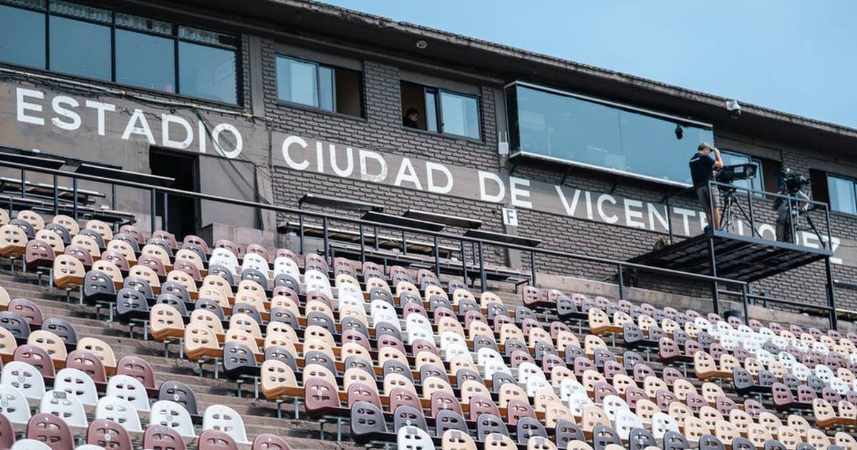 Empty stadium seats with a view of Estadio Ciudad de Vicente López sign, featuring filming equipment and operators.