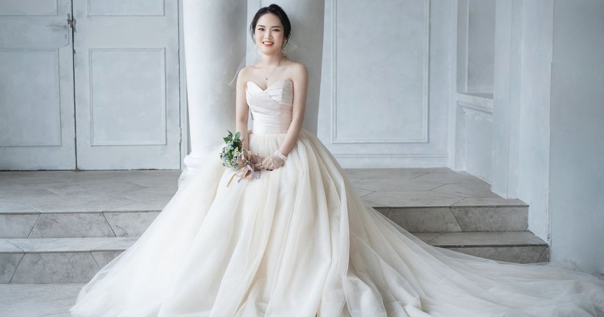 Beautiful bride seated indoors in a flowing wedding gown holding a small bouquet.