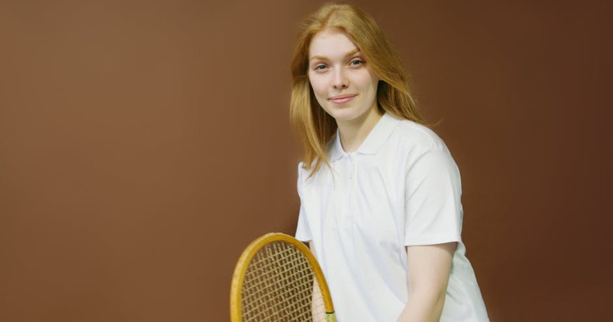 A smiling woman holds a tennis racket indoors, wearing a white shirt, ready for an athletic activity.