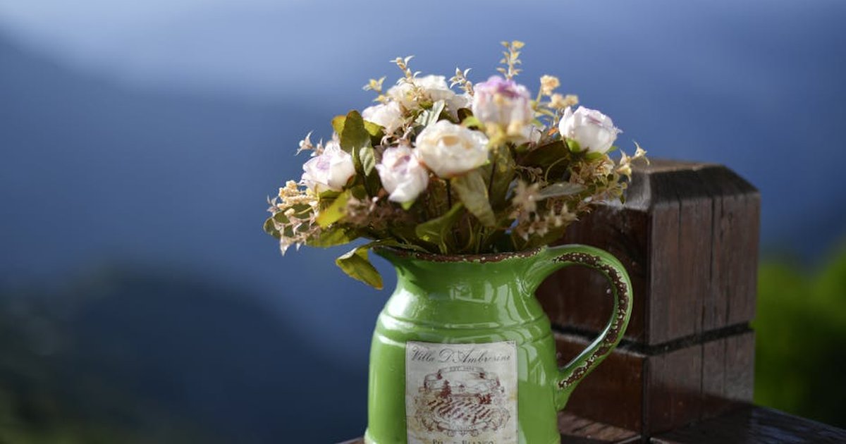 A green vase filled with roses and greenery sits on a wooden ledge against a mountainous backdrop.