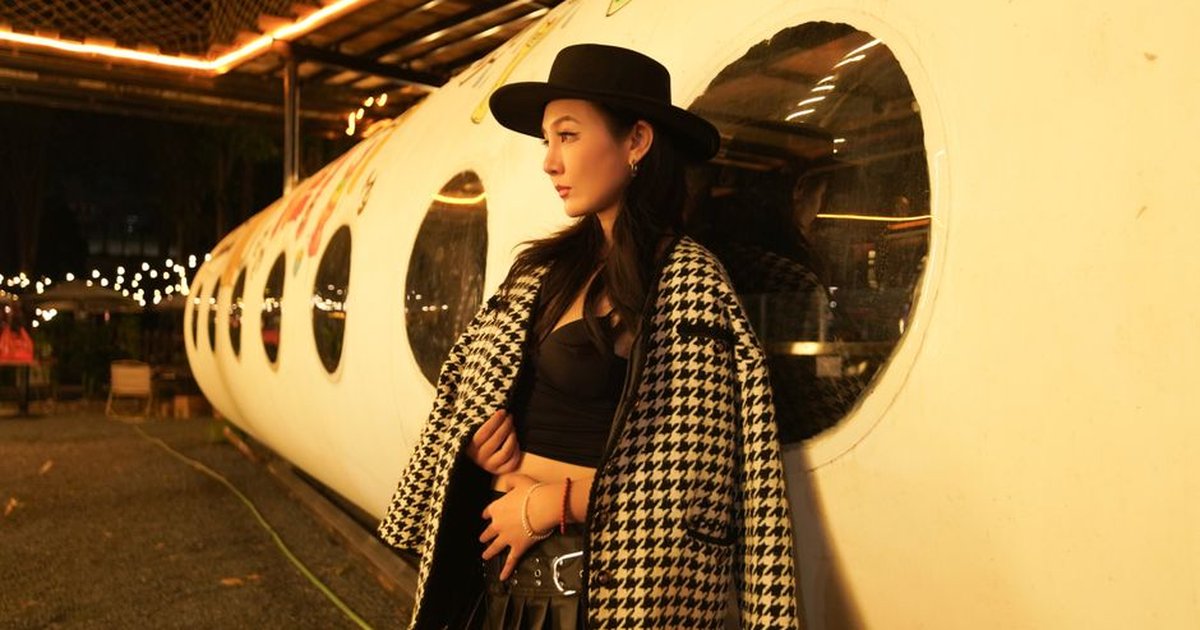 A fashionable woman in a houndstooth coat leans against a tube-shaped structure, illuminated by night lights.