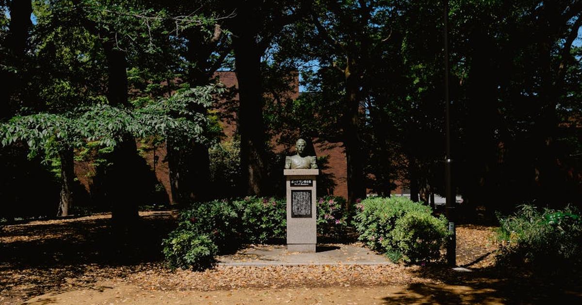 A serene sunlit park featuring a historical monument bust surrounded by lush greenery.