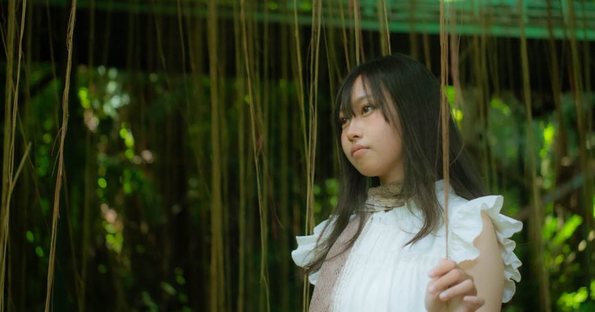 Portrait of a young woman in white dress surrounded by hanging vines in a lush forest setting.