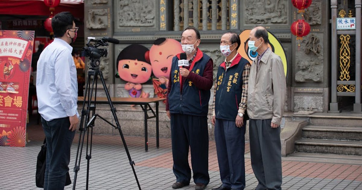 Elderly men wearing face masks being interviewed outdoors with festive decorations in the background.