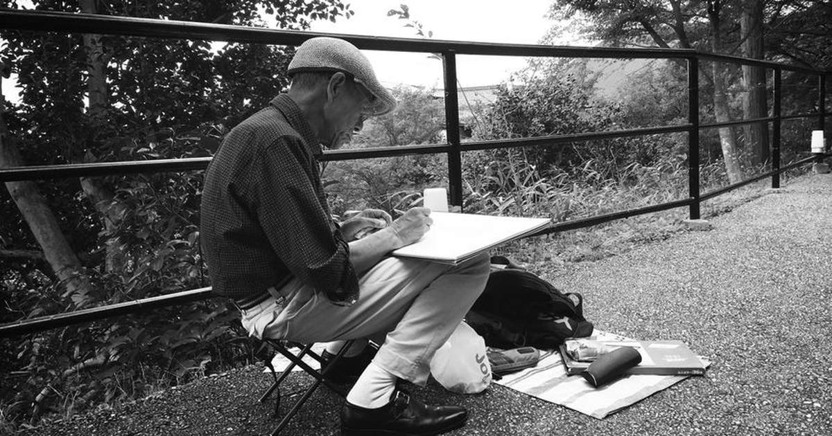 Black and white image of an elderly man sketching in a serene outdoor setting, embodying creativity and focus.