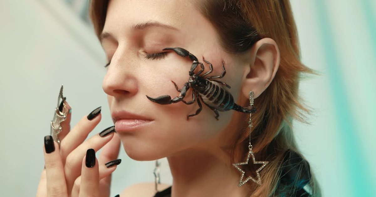 Close-up portrait of a woman with closed eyes, adorned with jewelry and a scorpion on her face.