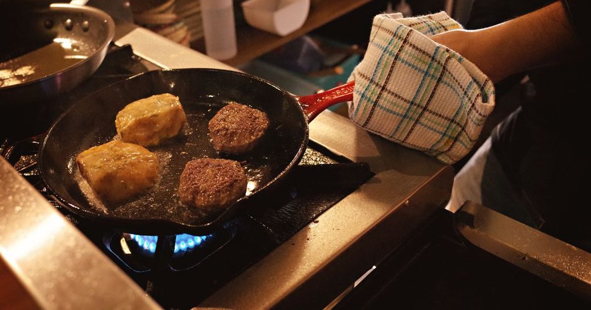 Close-up of two burgers and two patties cooking in a pan on a stove, kitchen scene.
