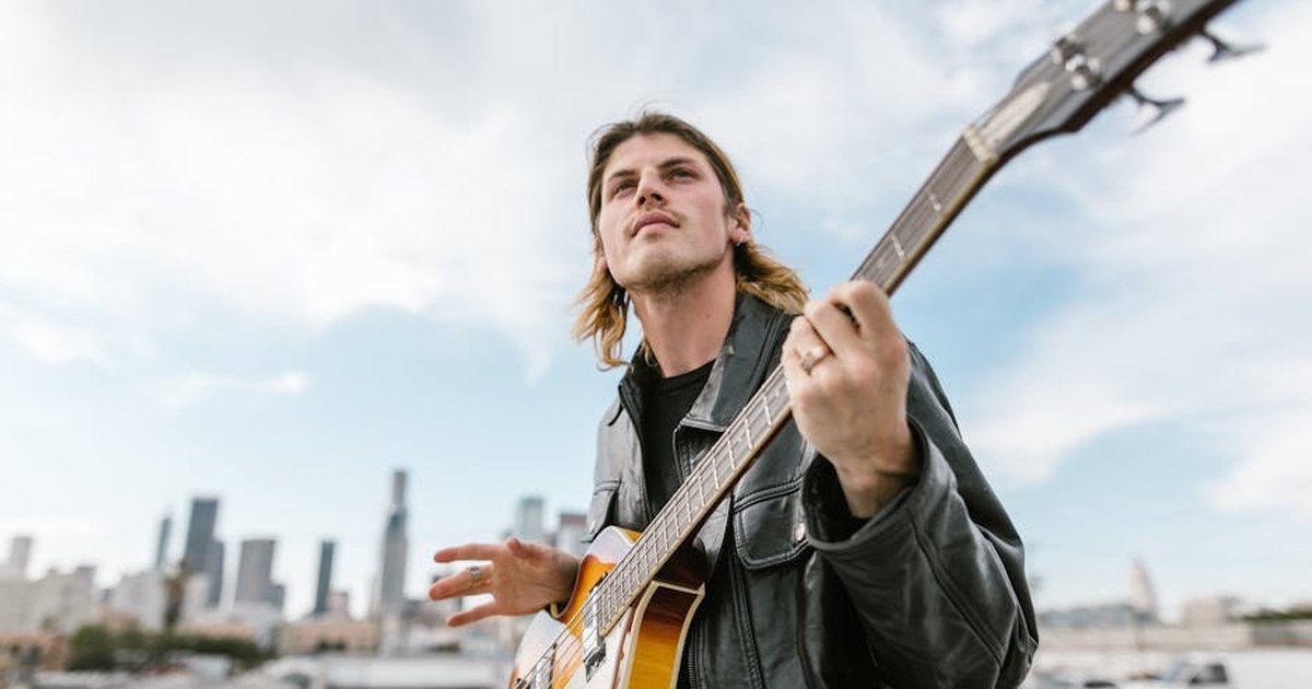 Caucasian musician in black leather jacket playing electric guitar on a rooftop with city skyline backdrop.