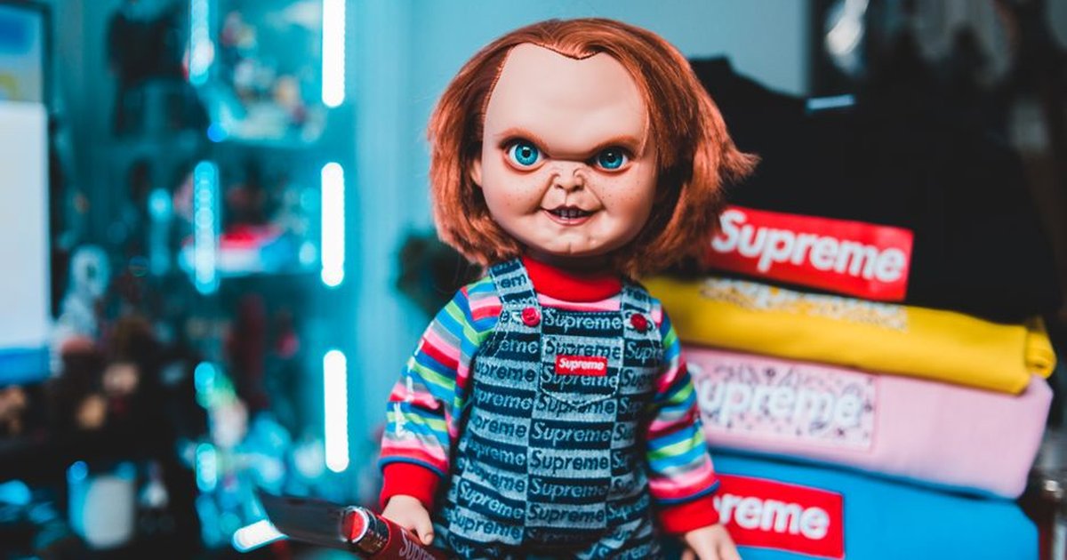 Scary Chucky doll wearing a Supreme outfit stands next to colorful Supreme clothing indoors.