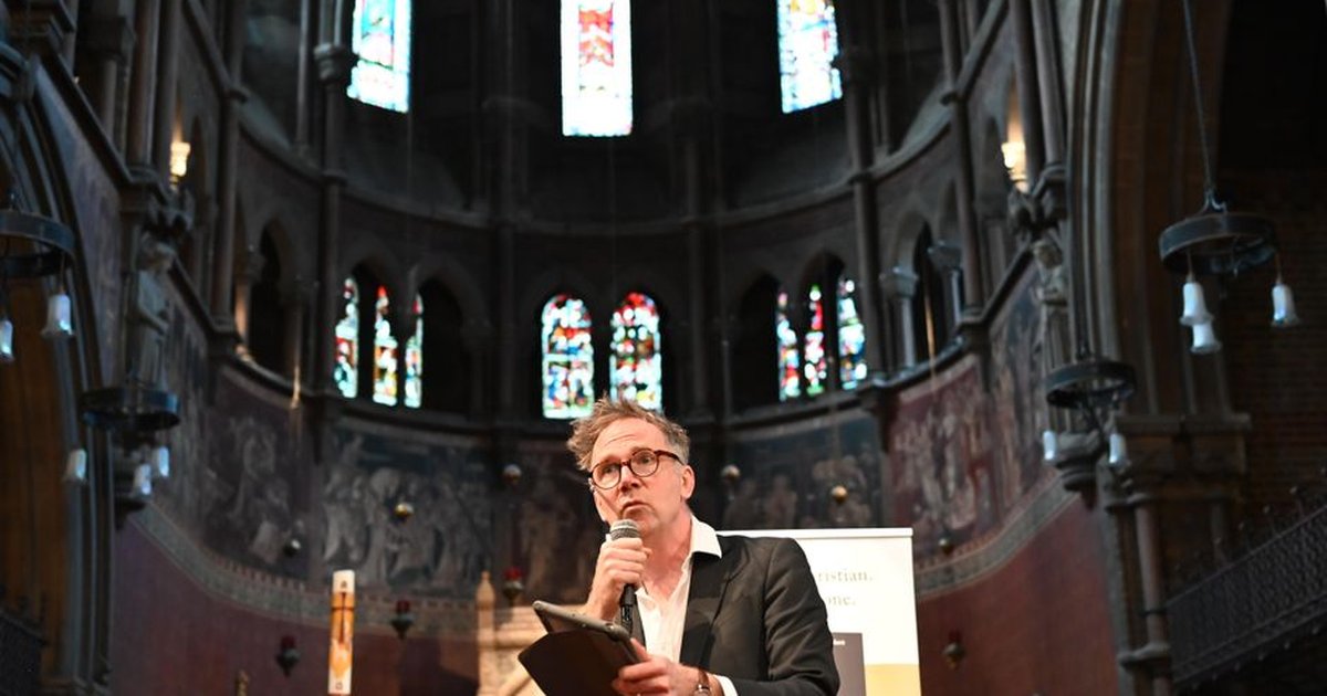 Man speaking in a historic church, surrounded by stained glass windows and ornate architecture.
