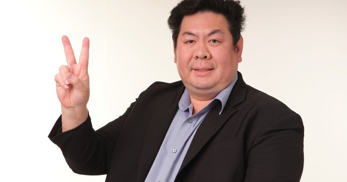 Smiling man in a suit showing peace sign against an indoor background.
