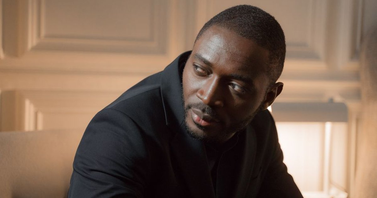 Thoughtful man in a black suit seated in a warmly lit indoor setting, reflecting an elegant mood.
