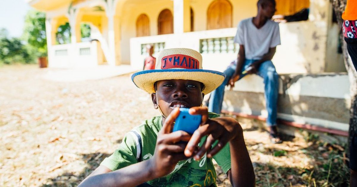 A young boy enjoys leisure time outdoors in Haiti, wearing a straw hat and holding a phone.