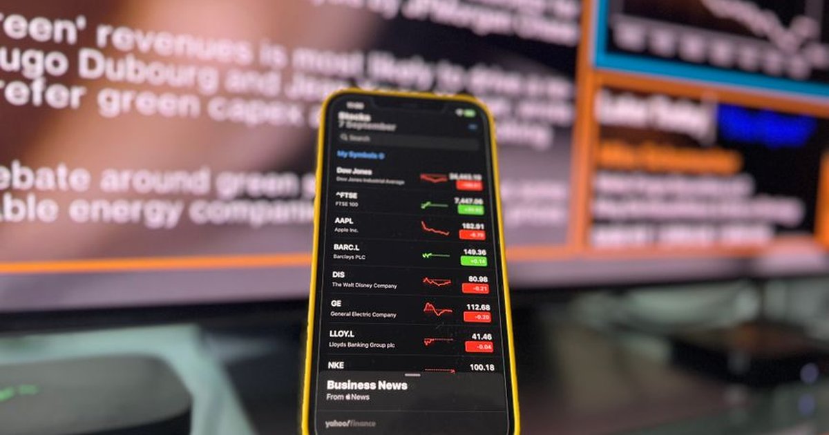Smartphone showing stock market trends in front of blurred financial news screen indoors.