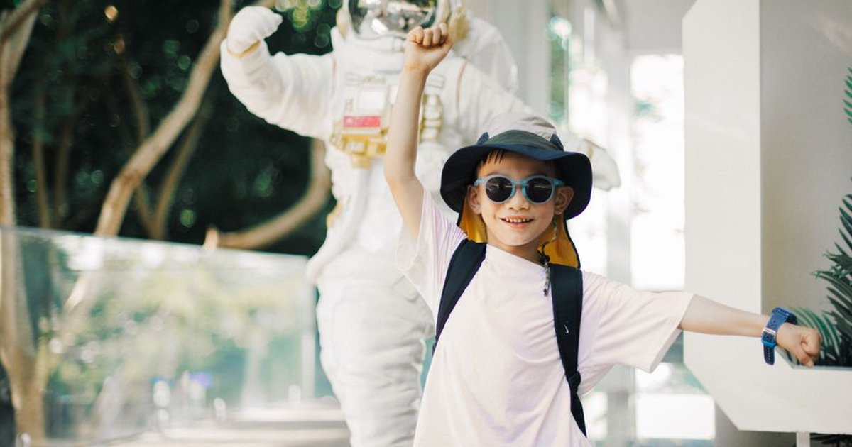 A smiling child in a hat and sunglasses posing beside an astronaut statue in an outdoor setting.
