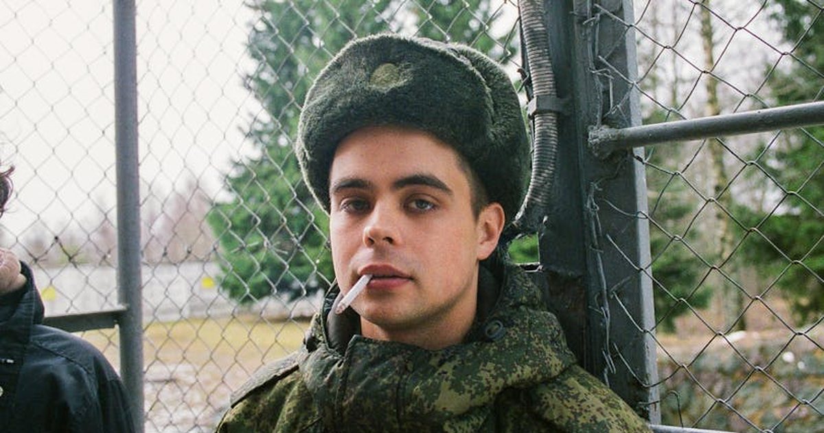 A young soldier in camouflage uniform smokes a cigarette by a chain-link fence in a winter setting.
